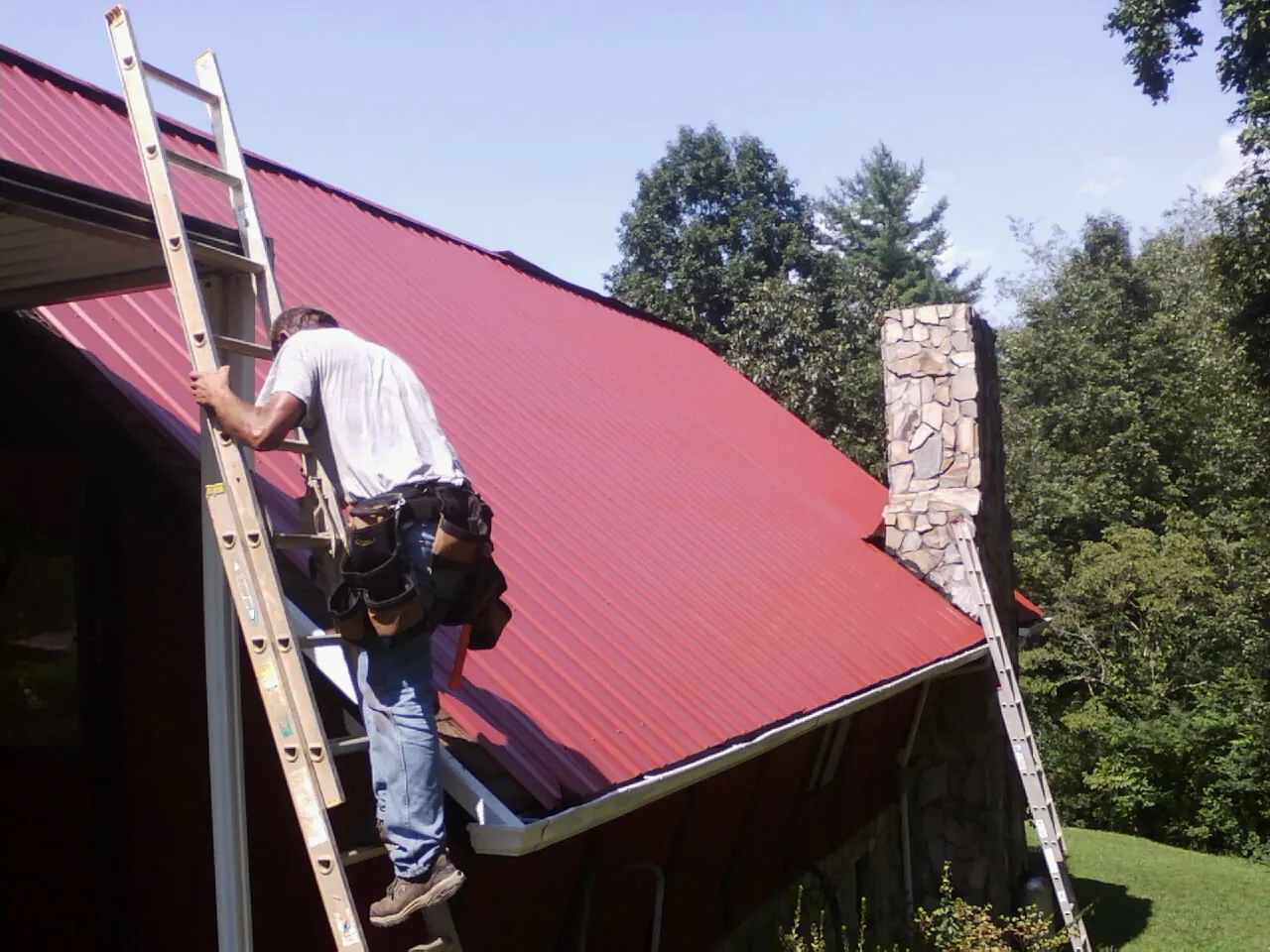Roofer on a ladder beside a red metal roof and stone chimney — Soffit & Fascia Repair by Roofing Brothers in Leonidas